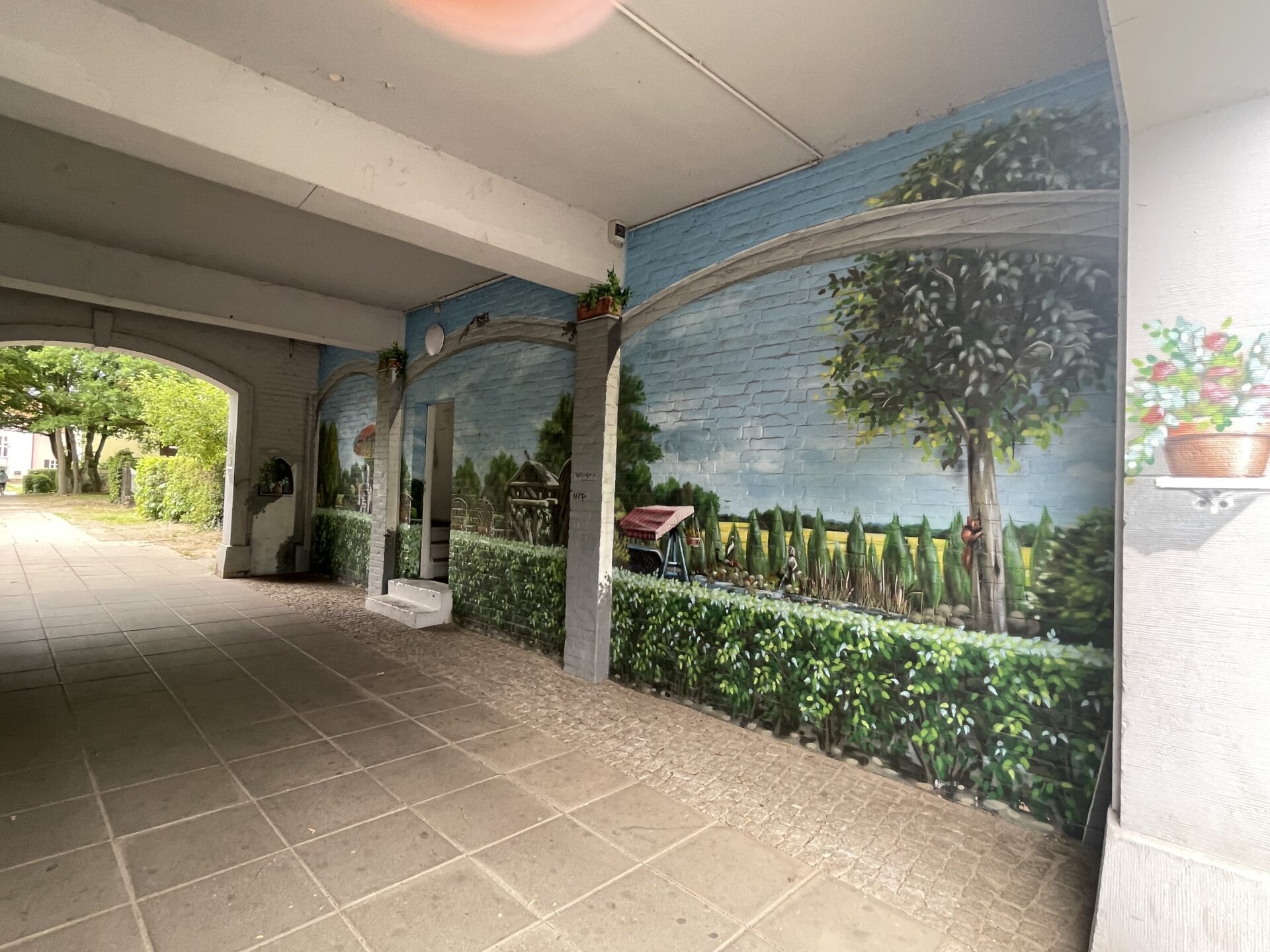 Überdachter Außenbereich mit Wandmalerei in Gartenoptik vor dem Friseursalon.