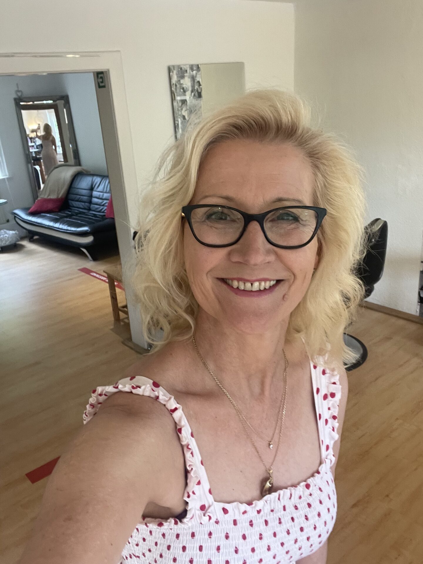 Porträt einer lächelnden Frau mit blonden Haaren und Brille im Friseursalon.