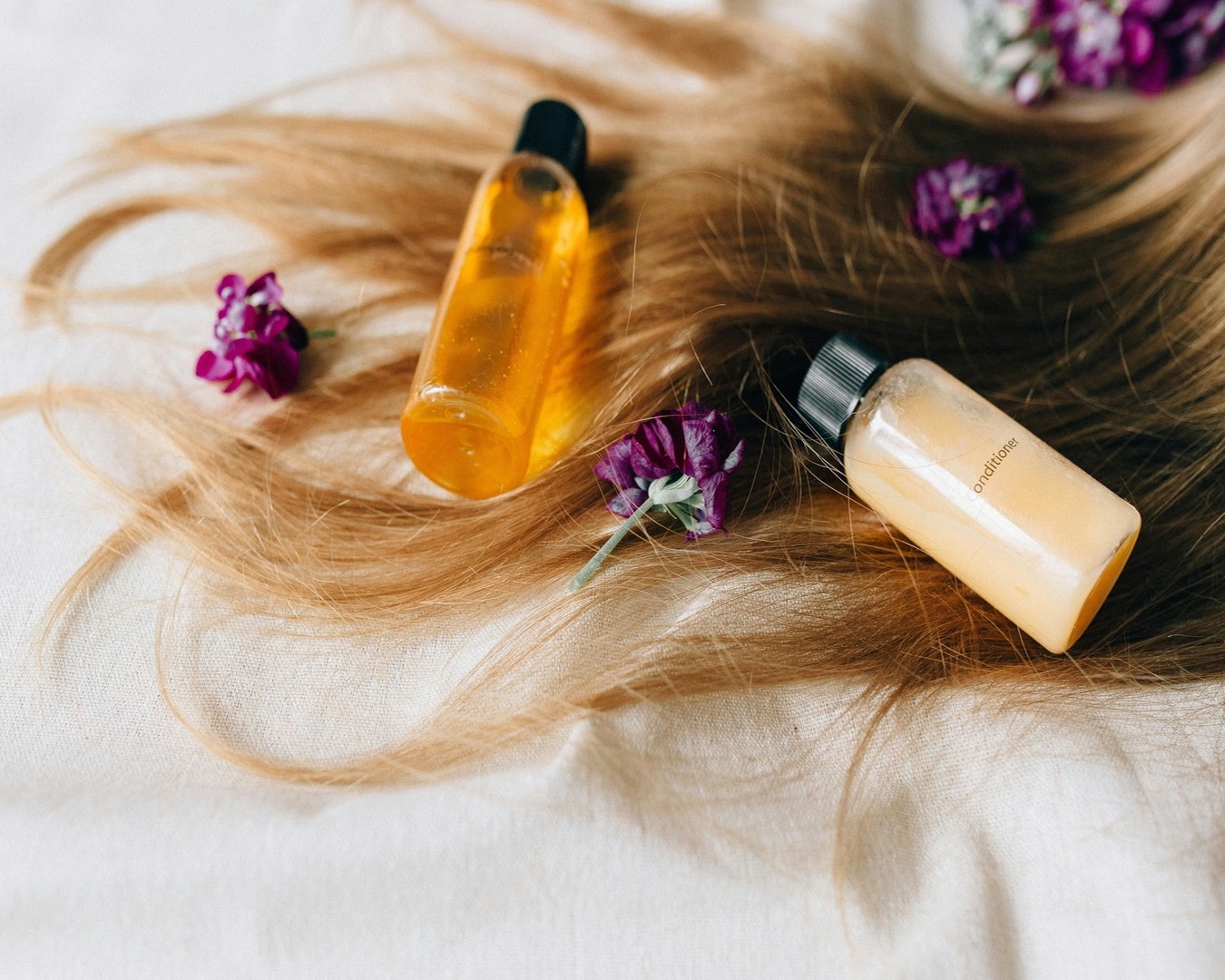 Blondes Haar auf weißem Hintergrund mit Violetten Blüten und zwei Flaschen Shampoo